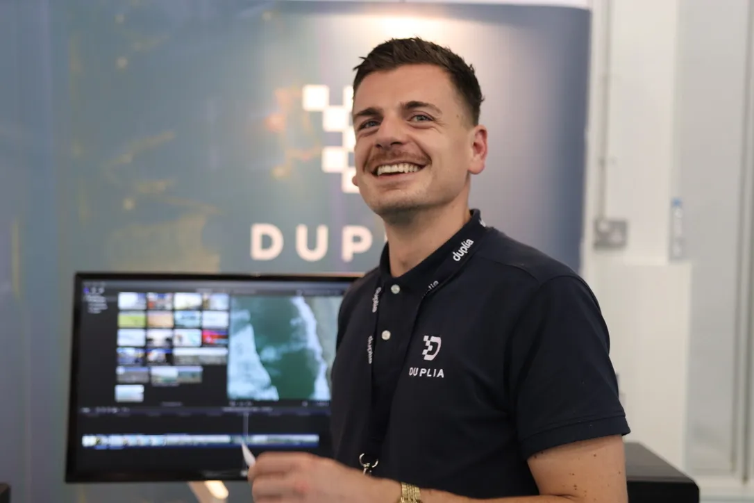 A smiling Duplia team member on the Duplia stand, beside an editing workstation